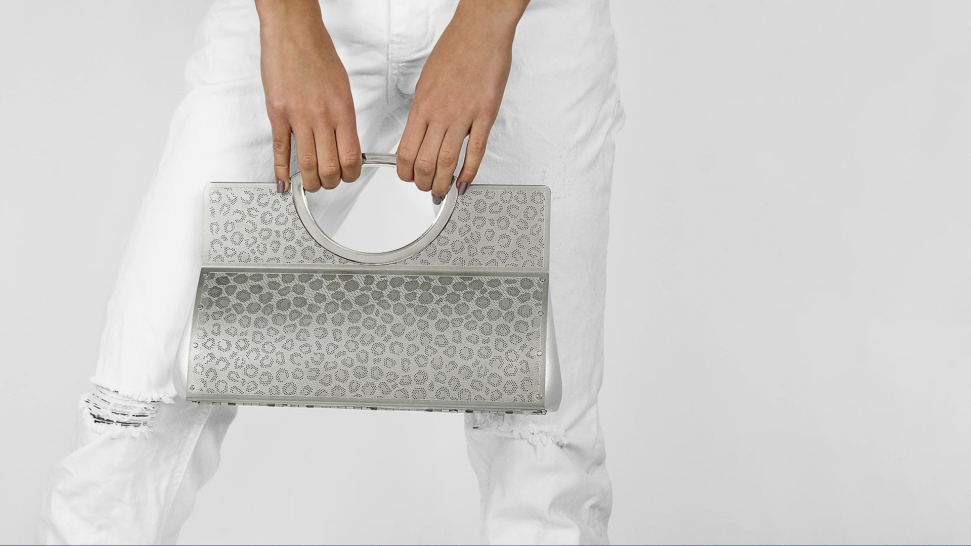 A person wearing white jeans holds a sleek, silver perforated metal handbag with a circular handle, showcasing modern design.