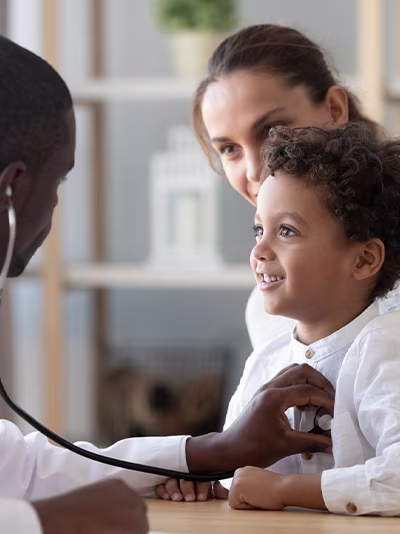 A doctor wearing a white coat and stethoscope listening to a child's heartbeat while mother looks on.