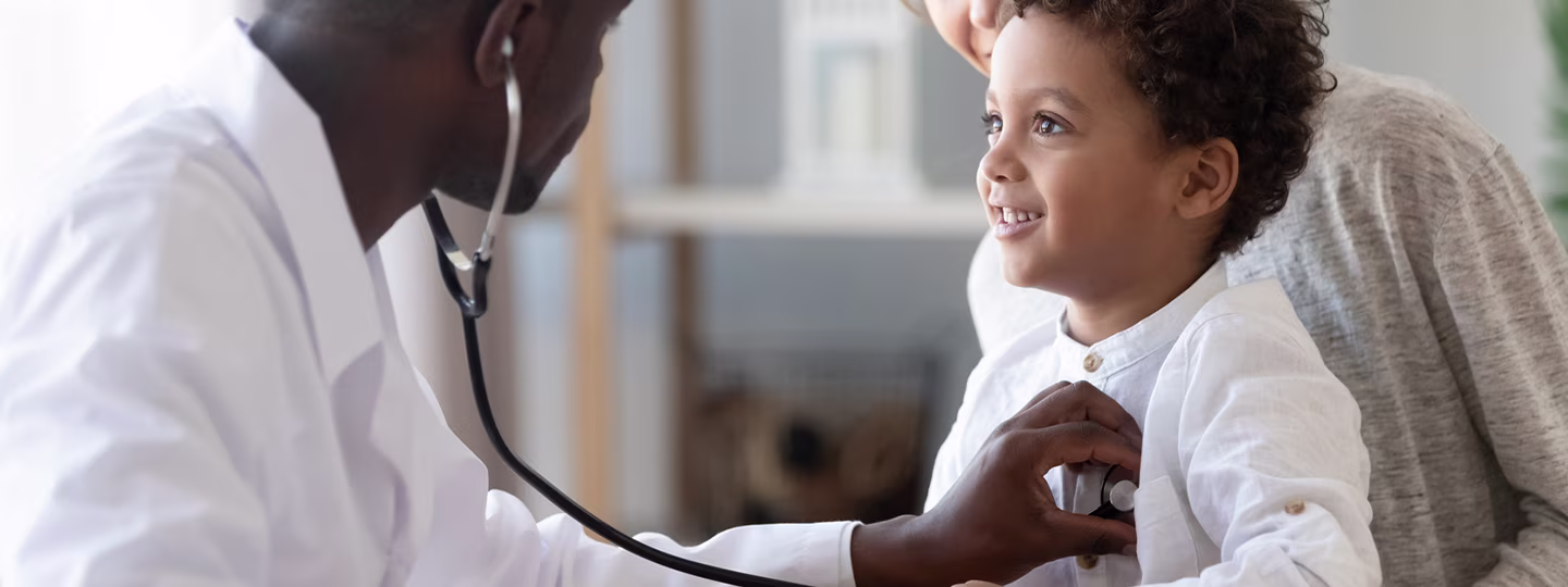 A doctor wearing a white coat and stethoscope listening to a child's heartbeat while mother looks on.