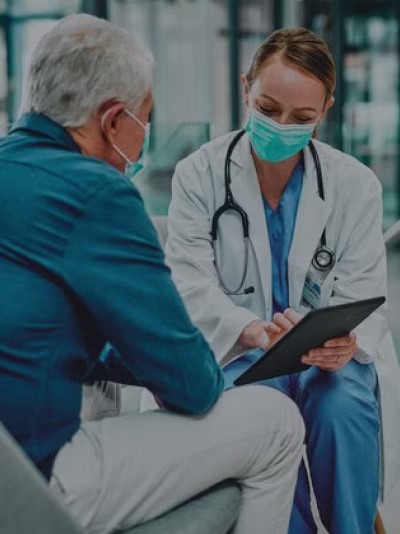 A lady Doctor wearing mask and white coat reviewing information on tablet with patient.