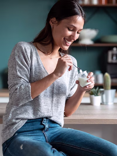 A woman eating a yogurt cup in a kitchen.