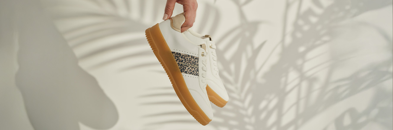 Main tenant des baskets blanches avec détail léopard et semelle en caoutchouc marron sur fond clair d'ombre de palmiers