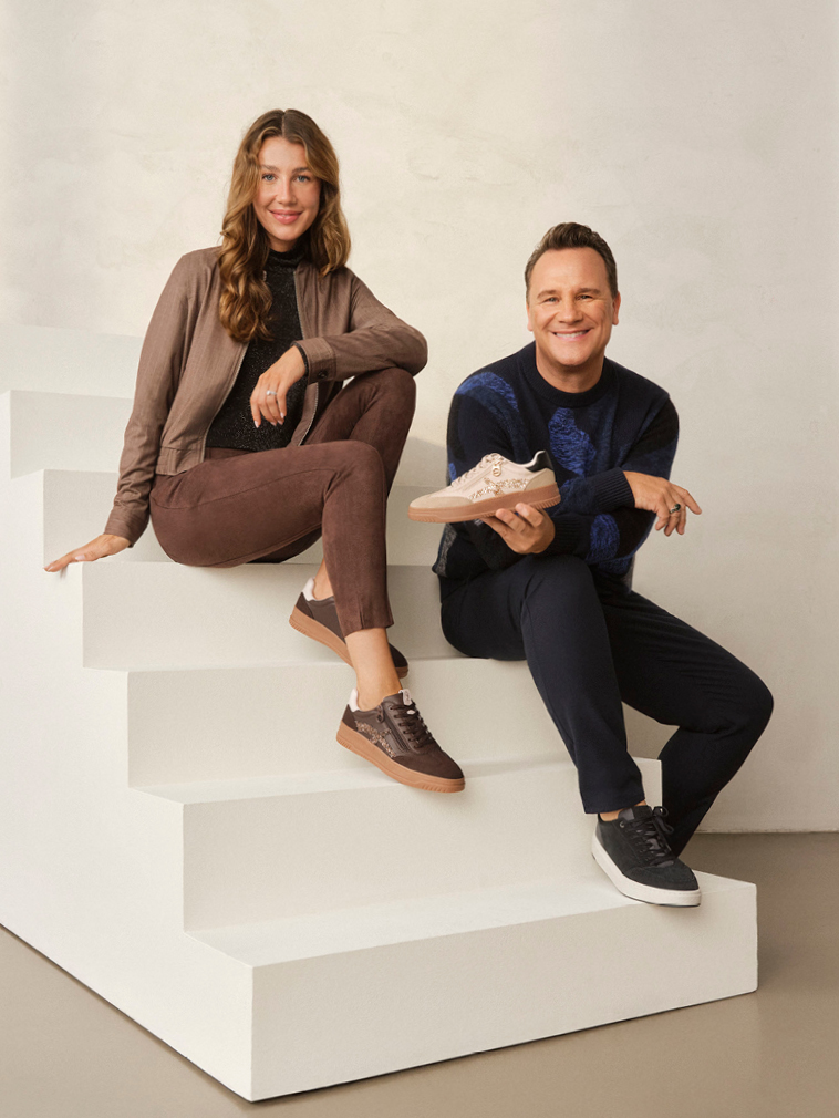 Frau in braunem Outfit und Mann in dunkler Kleidung sitzen lächelnd auf weißer Treppe und präsentieren Sneaker.