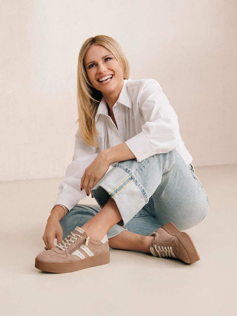 Lächelnde blonde Frau in weißer Bluse und Jeans sitzt auf dem Boden mit beigen Sneakers vor hellem Hintergrund.