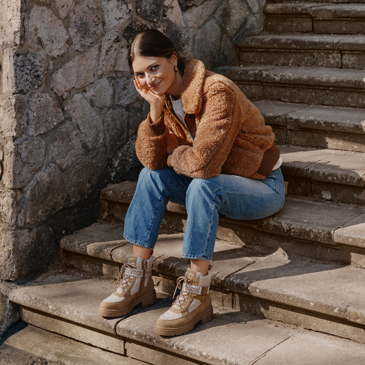 Lächelnde Frau in brauner Teddyjacke und Jeans sitzt auf Steintreppe vor alter Steinmauer.
