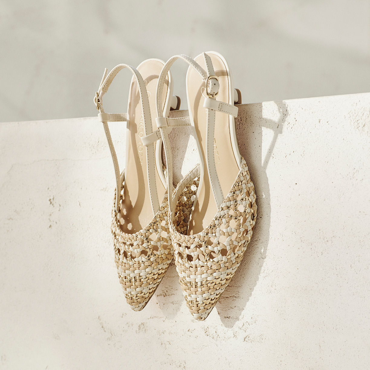 Scarpe slingback intrecciate beige con punta appuntita su una piattaforma di cemento bianco su uno sfondo neutro.