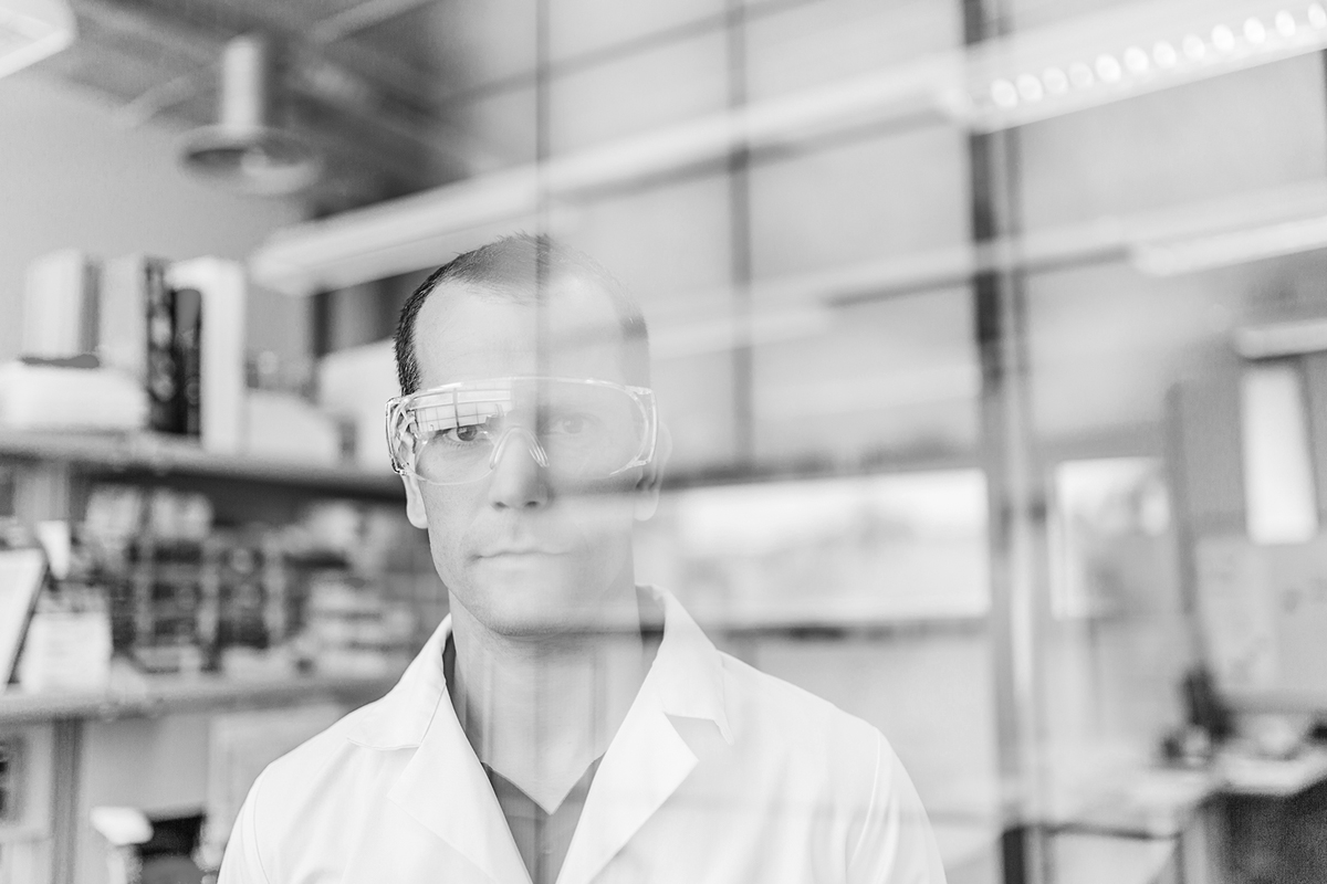 Black and white image of a scientist with safety glasses on staring into the camera from inside a lab