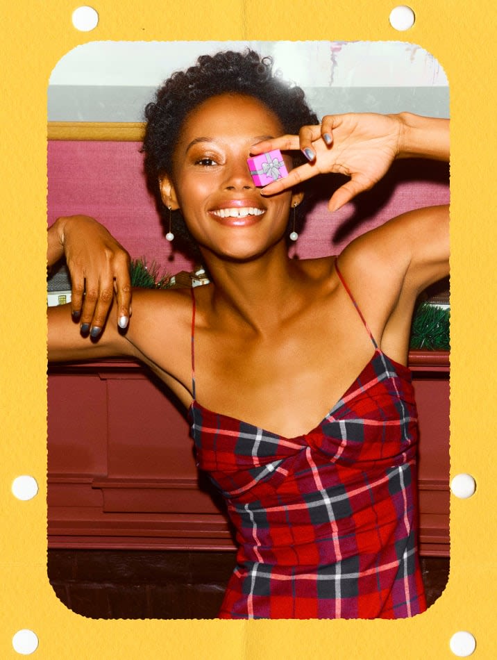 Model wearing a red tartan plaid slip dress, posing close-up with a tiny pink gift box over her eye.