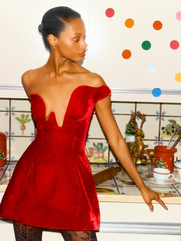 Model wearing a red velvet off-the-shoulder mini dress with a deep neckline, leaning against a kitchen counter.