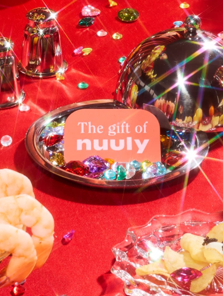 Pink Nuuly gift card presented under a silver cloche among jewels and party snacks on a red tablecloth.