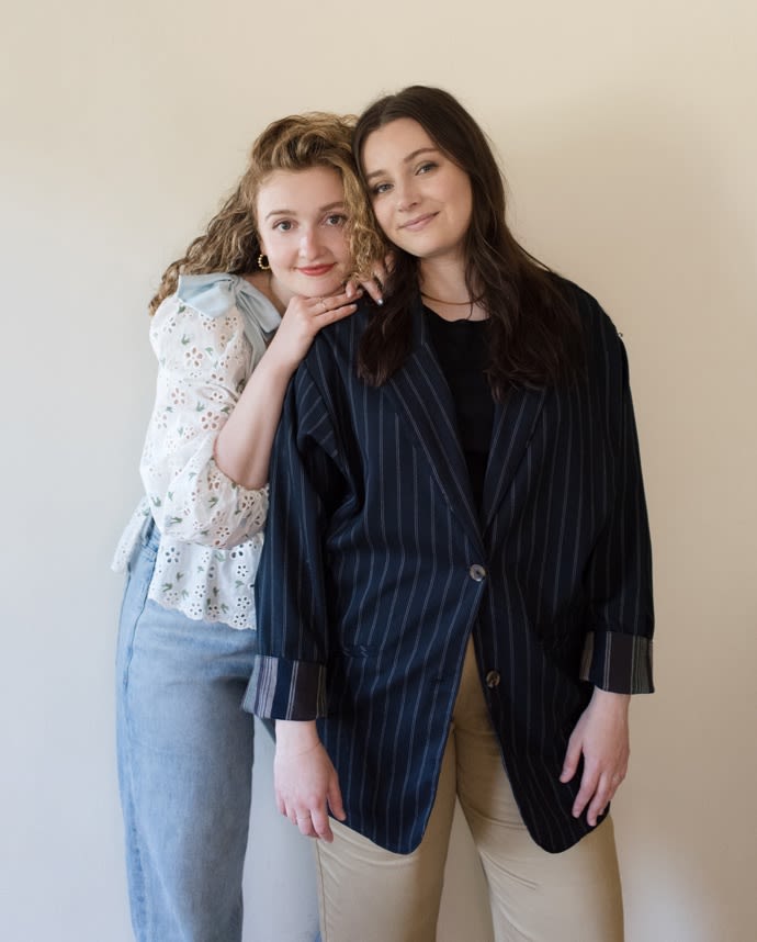 COUPLE HUGGING. GIRL ON THE LEFT WEARING JEANS AND A BLOUSE, AND A GIRL ON THE RIGHT WEARING PANTS AND A BLAZER.