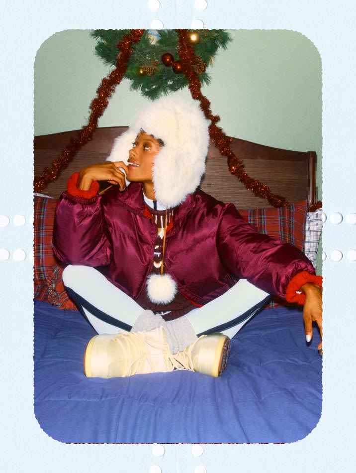 Model wearing a plum puffer jacket, white faux-fur trapper hat, and snow boots, cross-legged on a festive bed.