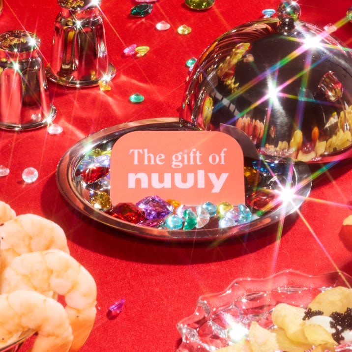 Pink Nuuly gift card presented under a silver cloche among jewels and party snacks on a red tablecloth.