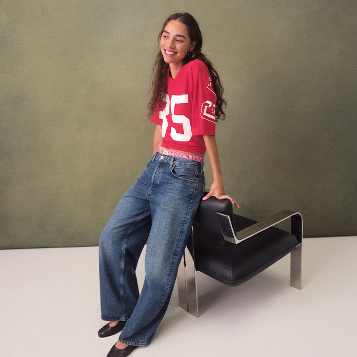 Model leans against a chair, styled in an over-sized red and white varsity-style jersey, barrel jeans, and black flats.