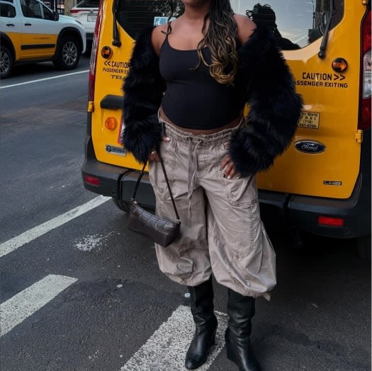 Model wearing a black faux-fur shrug, cropped tank, and cargo pants, standing behind a yellow city cab.