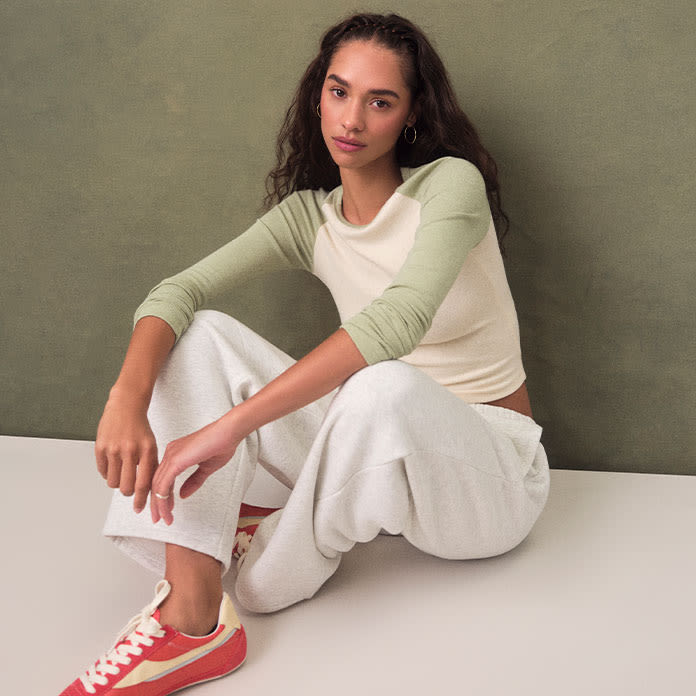 Woman sits in baseball long sleeve, jersey cream sweatpants and statement red sneakers.