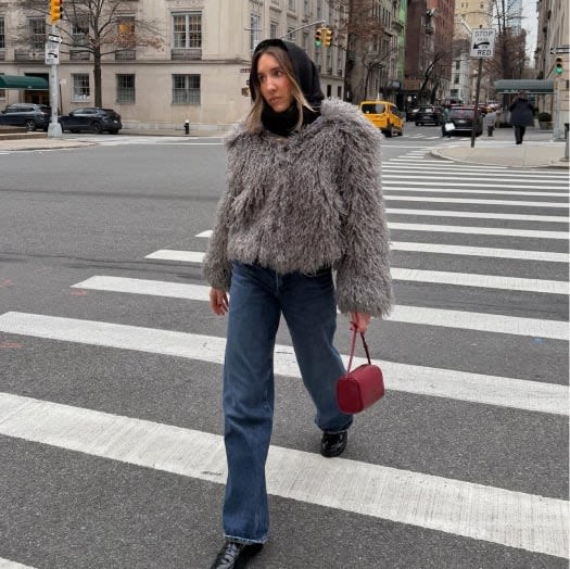 Model wearing a shaggy grey faux-fur coat, dark hoodie, and straight-leg jeans, walking across a crosswalk.