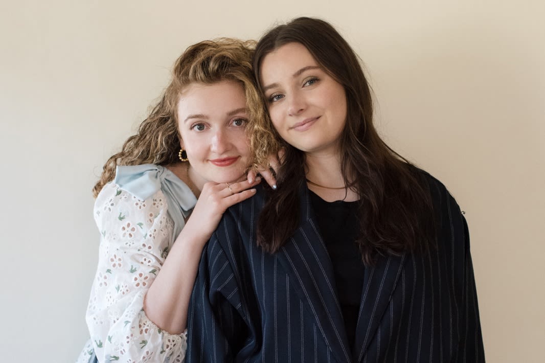 TWO GIRLS HUGGING. ONE GIRL WEARING A DRESS AND ANOTHER GILR WEARING A BLAZER.