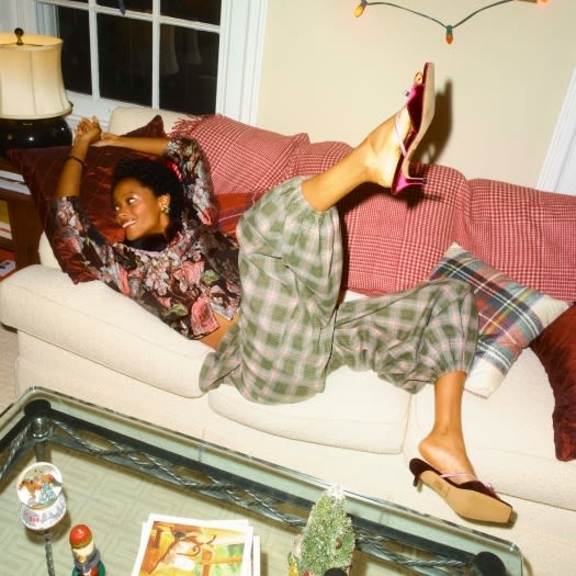 Model in a floral blouse and green plaid trousers lounging on a sofa, kicking up a pink velvet heel.