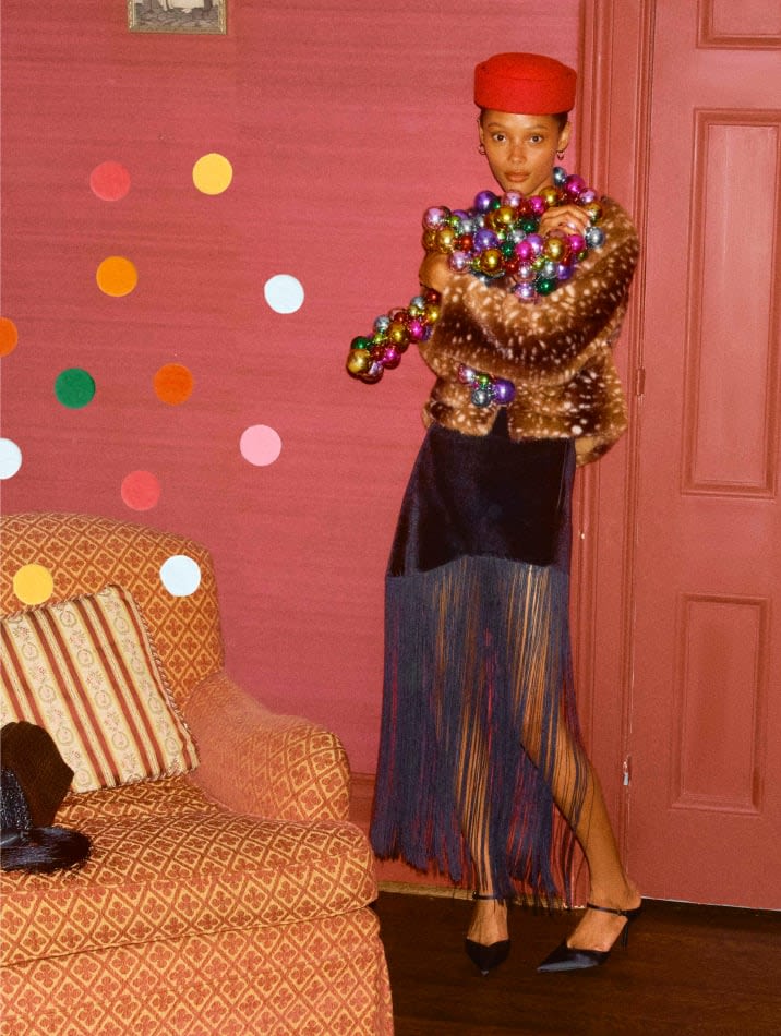 Model wearing a spotted faux-fur coat, fringe skirt, and red pillbox hat, draped in colorful ornament garland.
