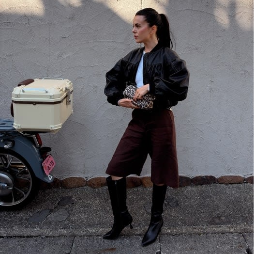 Model wearing a black cropped bomber jacket, tailored culottes, and tall boots, standing beside a scooter.