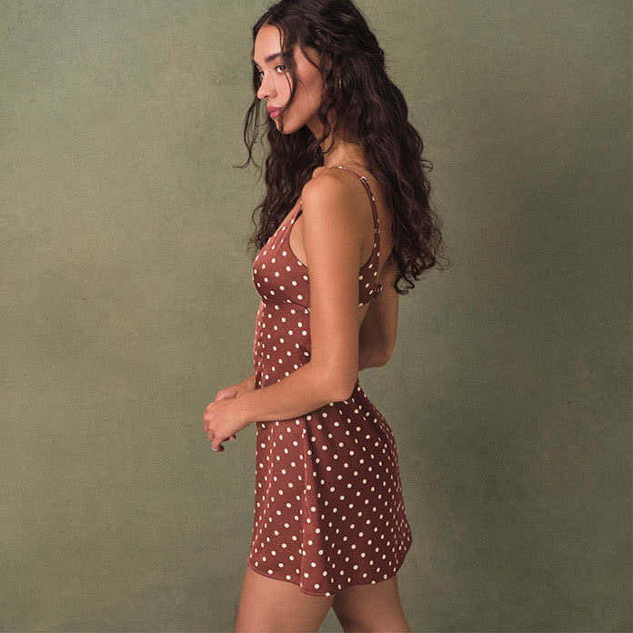 Model strikes shoulder in brown and white polka-dot mini dress with small straps and an open back.