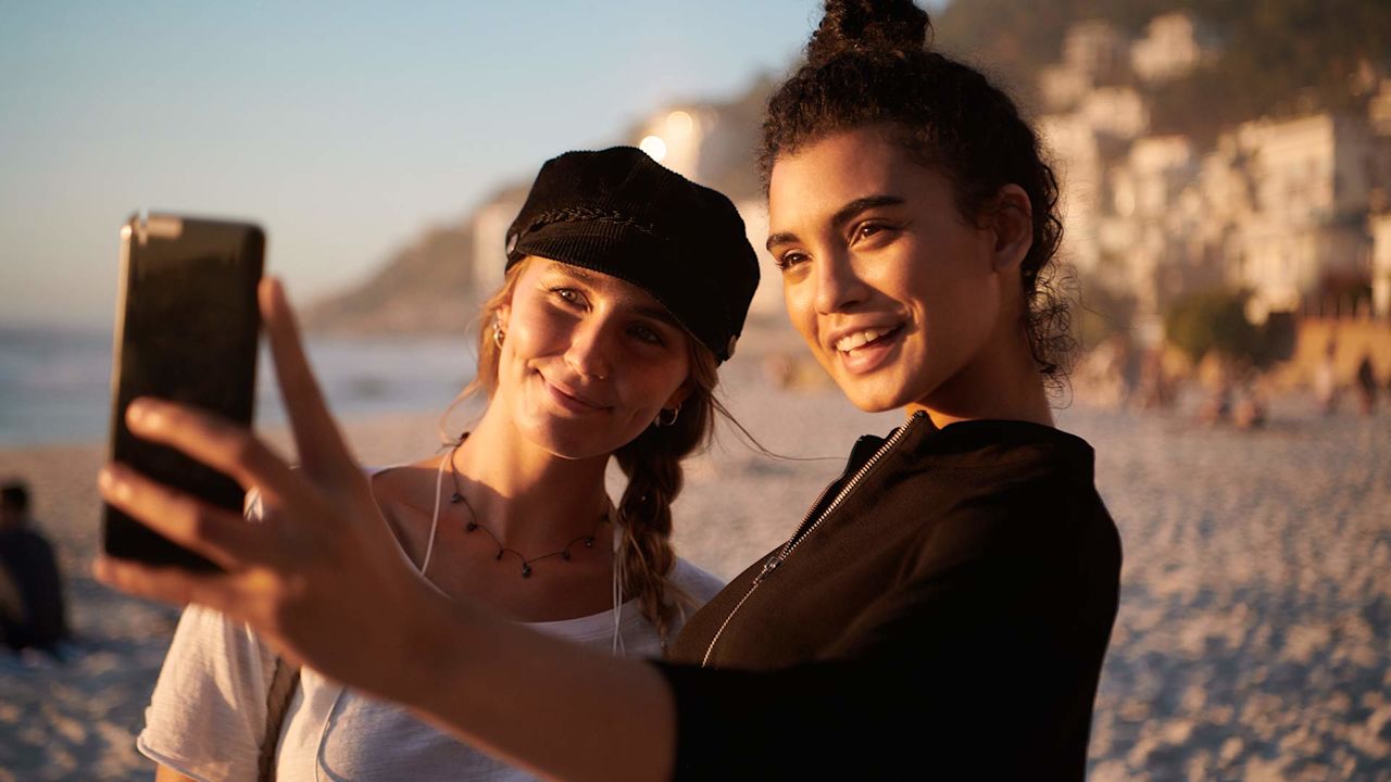 Två kvinnor vid havet tar en selfie med kvällssolen i ansiktet.