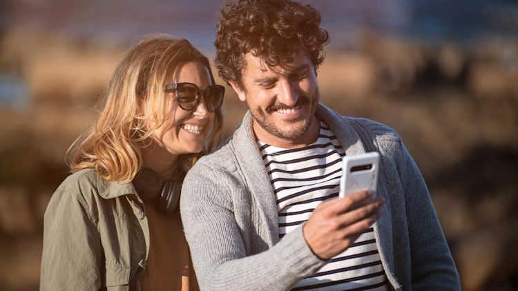 Kvinna och man. Kvinnan tittar över mannens axel. Mannen håller i en mobiltelefon.