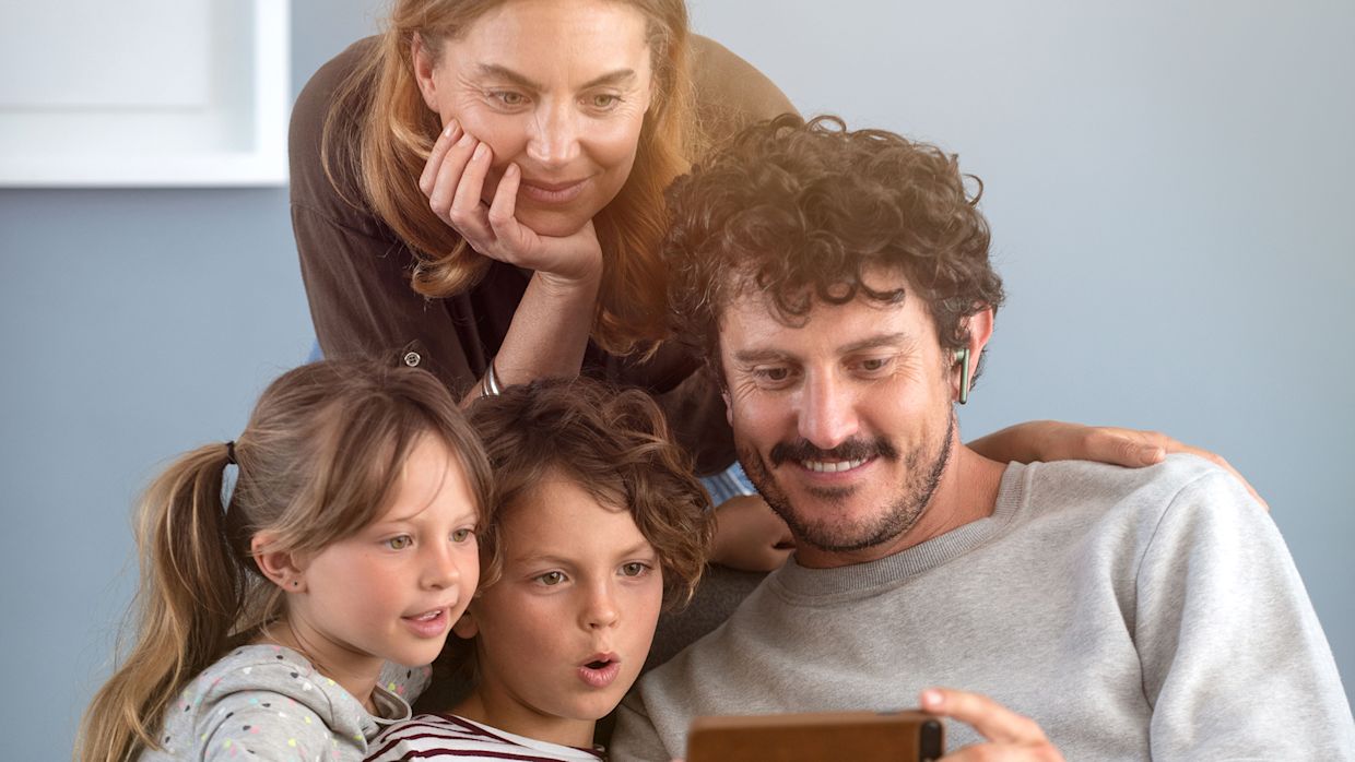 En familj sitter tätt tillsammans och tittar på mobiltelefonen som pappan håller i handen.