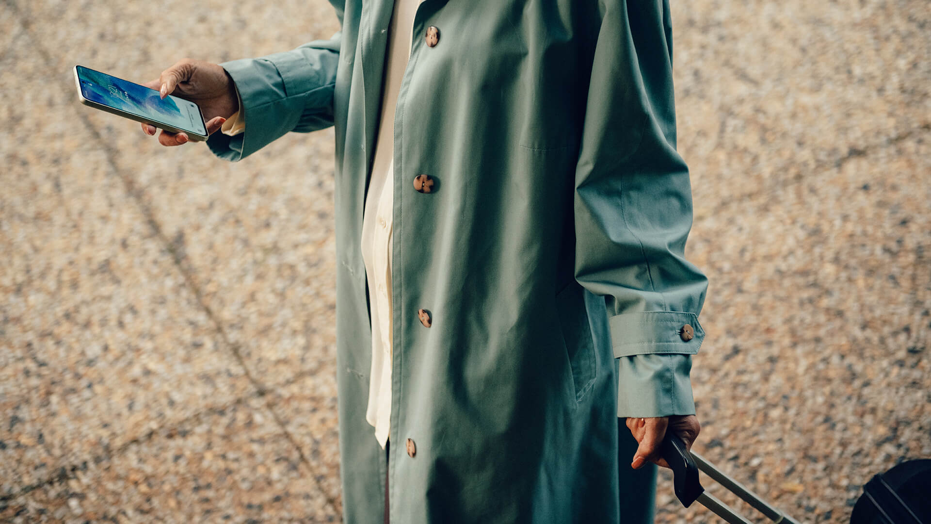 En kvinna i grön trenchcoat scrollar på sin mobiltelefon medan hon går med en flygväska i handen.