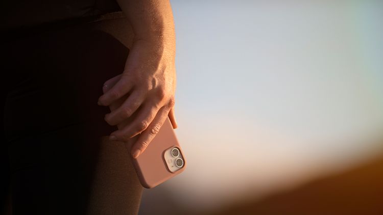 Hand hållandes en mobiltelefon som pekar neråt. Mobilen har ett skal i rostrosa färg. Foto är taget i midjehöjd.