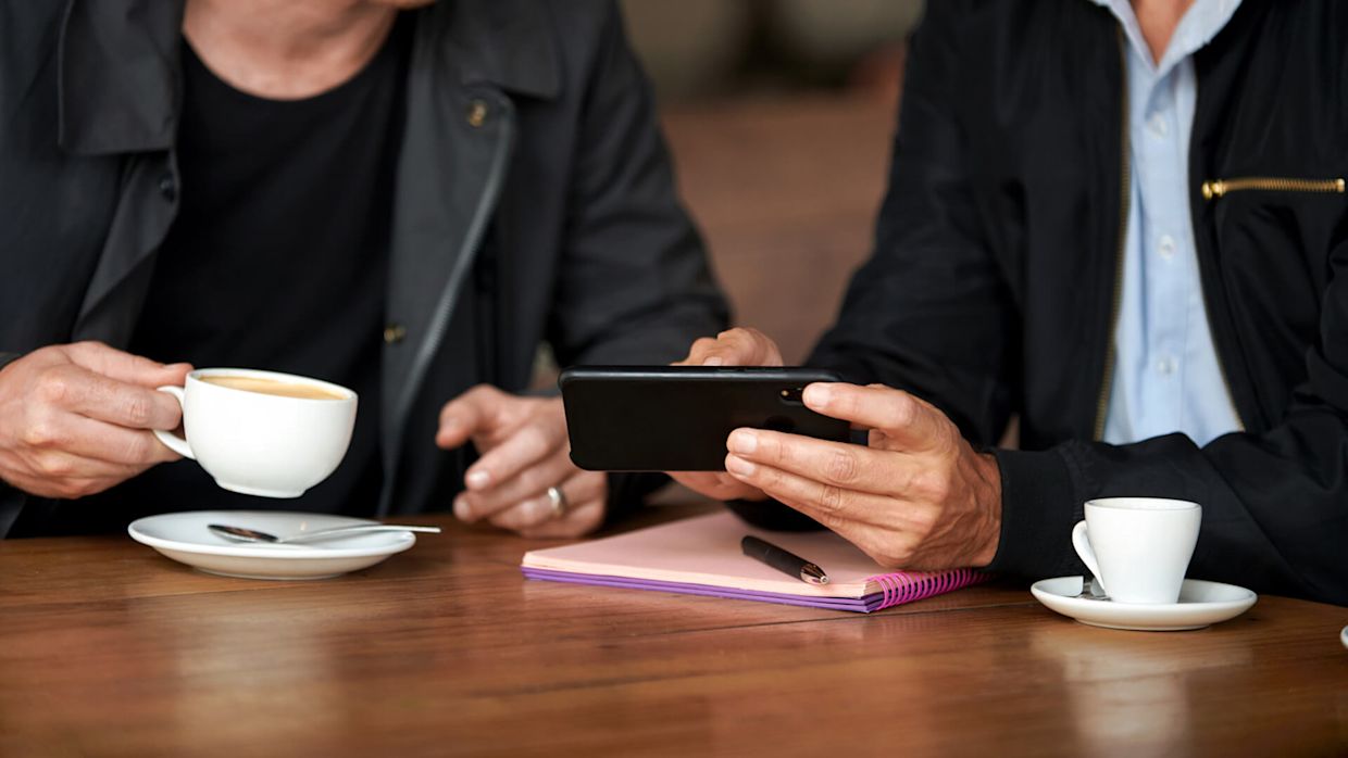 Två personer i mörka kläder sittandes vid ett bord med varsin kaffekop. En av personerna visar den andra något på en mobiltelefon.