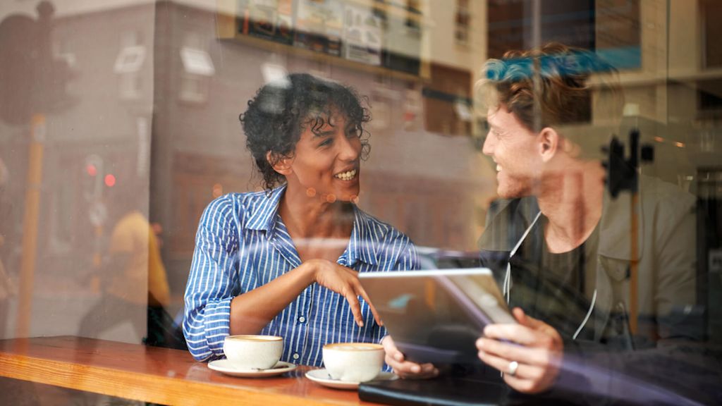 Man och kvinna vid ett bord på ett cafè med varsin kaffekopp. De pratar. Mannen håller i en surfplatta.
