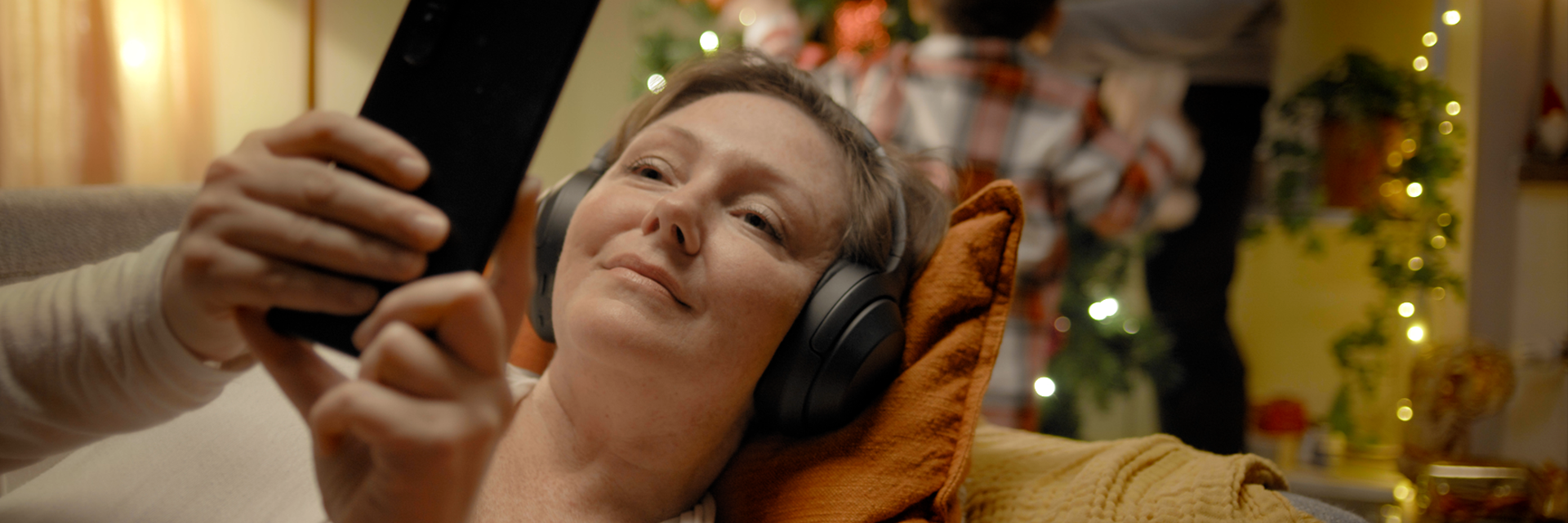 Kvinna som ligger i soffan och lyssnar på musik i hörlurarna och tittar på mobilen