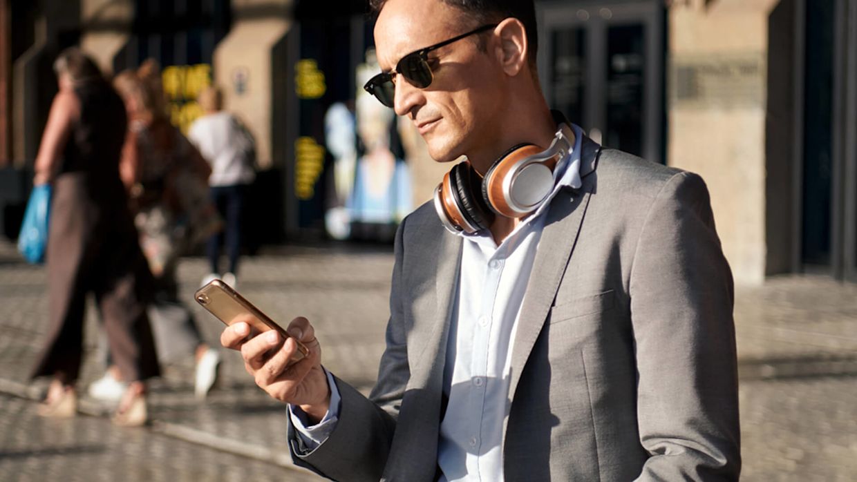 Man i grå kavaj, svarta solglasögon och hörlurar runt halsen hållandes i en mobil i ena handen.