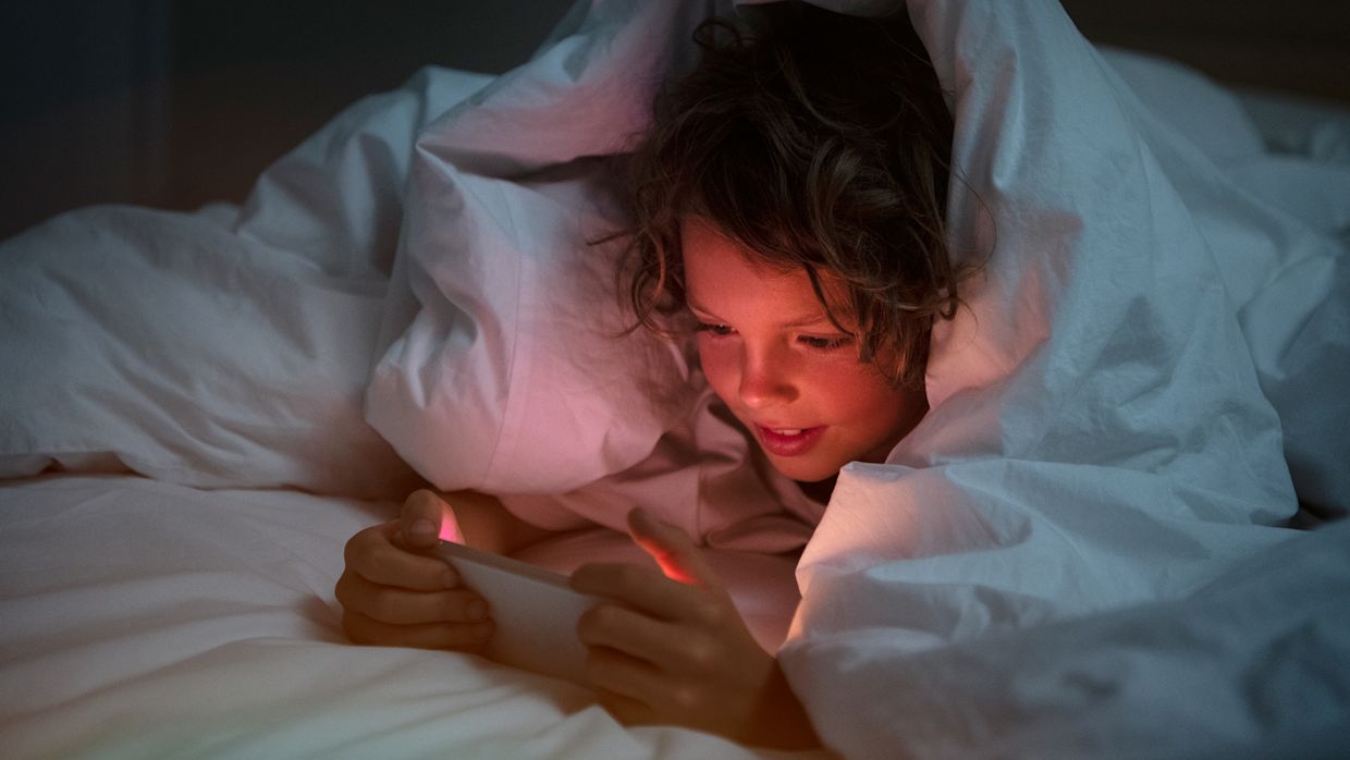 Barn som ligger under ett täcke i en säng och tittar på något på mobilen.