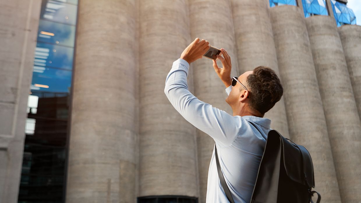 Kortklippt man i ljusblå skjorta, svart ryggsäck och solglasögon fotandes en grå betong-byggnad.