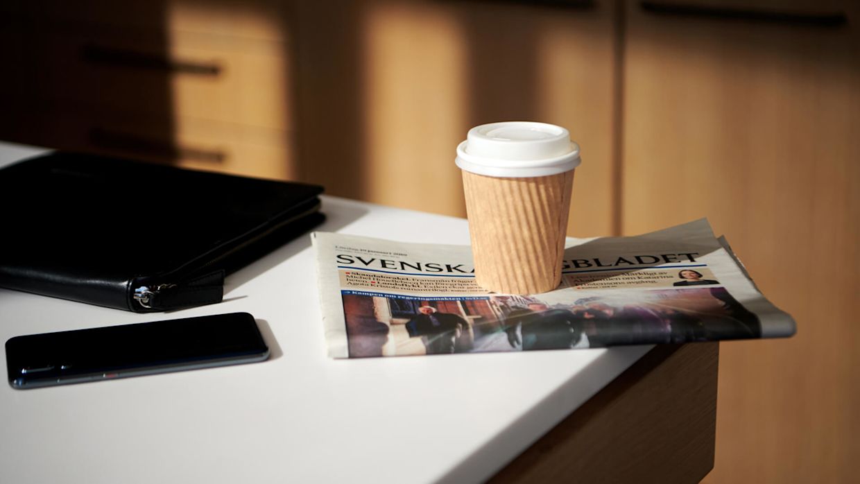 Ett vitt skrivbord med en mobiltelefon, surfplatta, tidning och en kopp kaffe