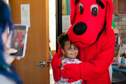 A child hugging Clifford the Big Red Dog while an adult, in the foreground, takes a picture on a smart phone.