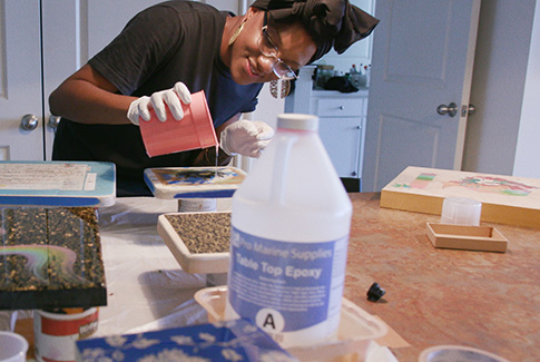 A woman, wearing gloves, pouring liquid from a cup as part of an at-home craft project. There is a bottle labeled Table Top Epoxy in the foreground.