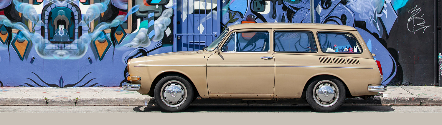 A tan colored two door station wagon from the 70's parked in front of a blue and black wall mural..