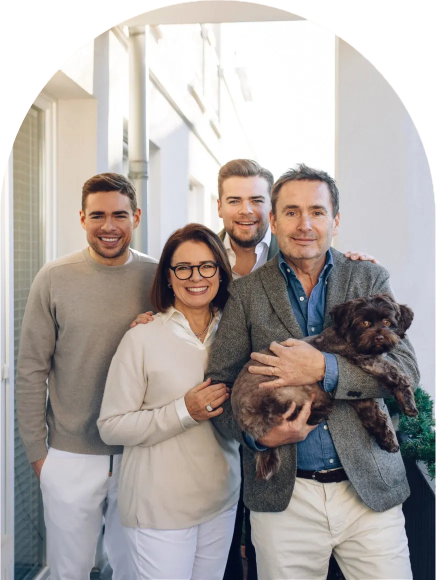 Eine lächelnde vierköpfige Familie, darunter zwei Männer, eine Frau und ein älterer Mann, der einen kleinen braunen Hund hält, steht eng beieinander in der Nähe eines weißen Torbogens.