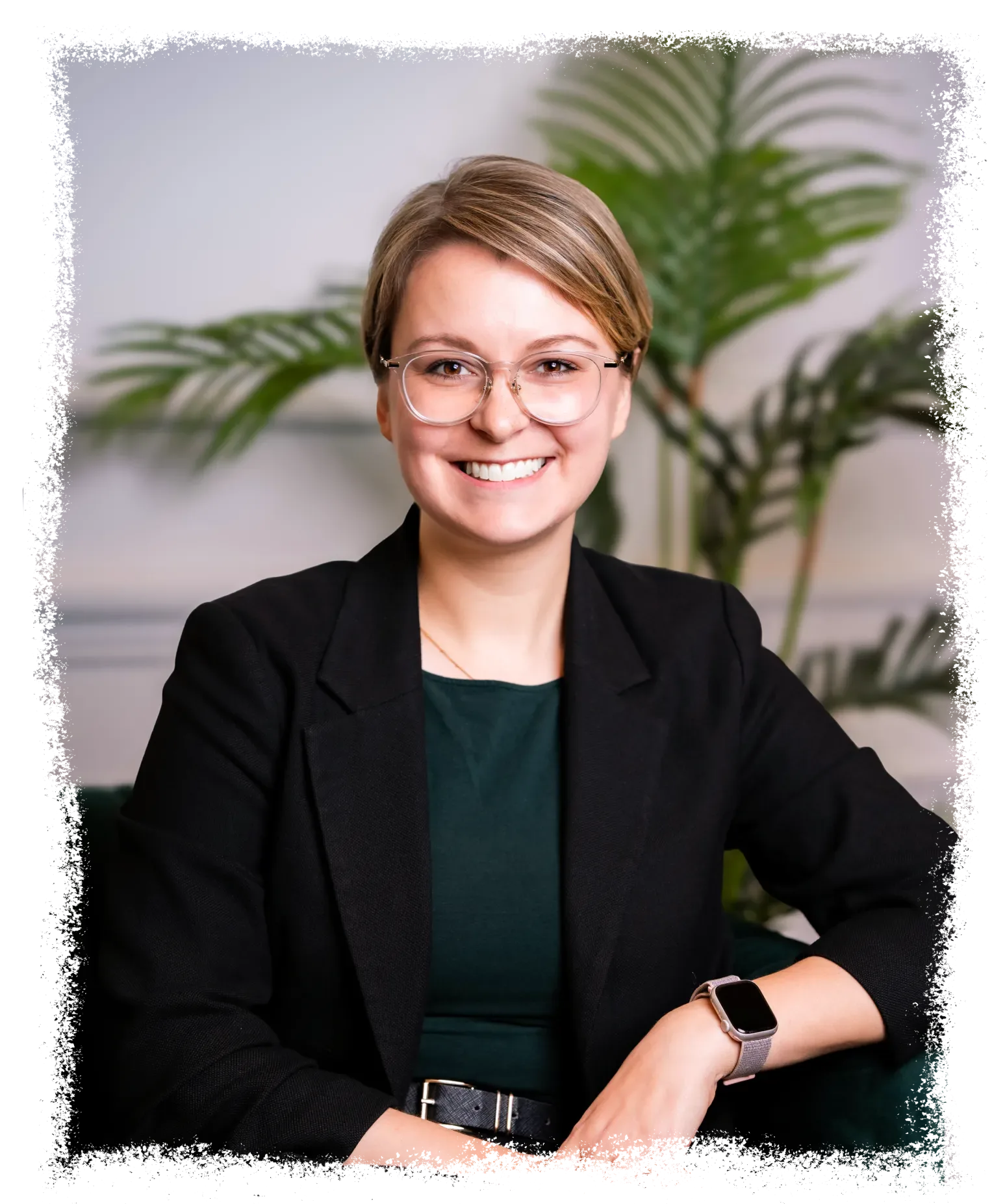 Man sieht die Zitatgeberin Caroline Nix von rimc. Mit kurzen blonden Haaren und Brille sitzt in einem dunklen Blazer und einem grünen Oberteil und trägt eine Smartwatch. Im Hintergrund ist eine Blattpflanze zu sehen. Das Bild hat einen weichen, weißen Randeffekt.