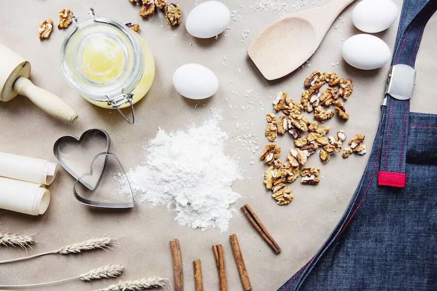 Baking ingredients and tools, including eggs, walnuts, flour, cinnamon sticks, cookie cutters, a rolling pin, an apron, and a jar of honey, arranged on a table.