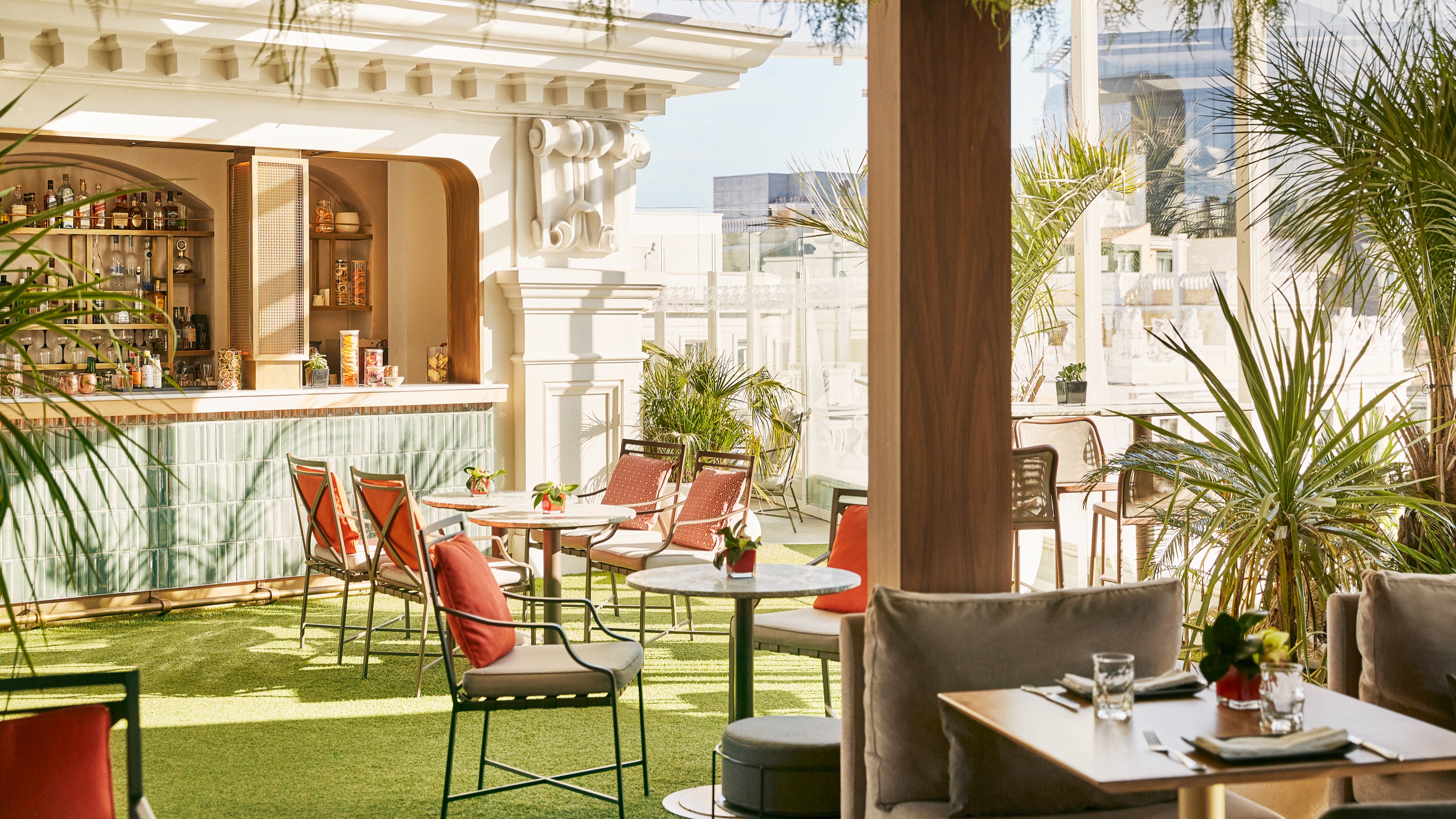 La imagen muestra la terraza del Jardin de Diana.Cuenta con una variedad de asientos, incluyendo mesas con sillas y sofás, todos dispuestos sobre césped artificial. En el fondo se puede ver el área del bar con estantes llenos de botellas.