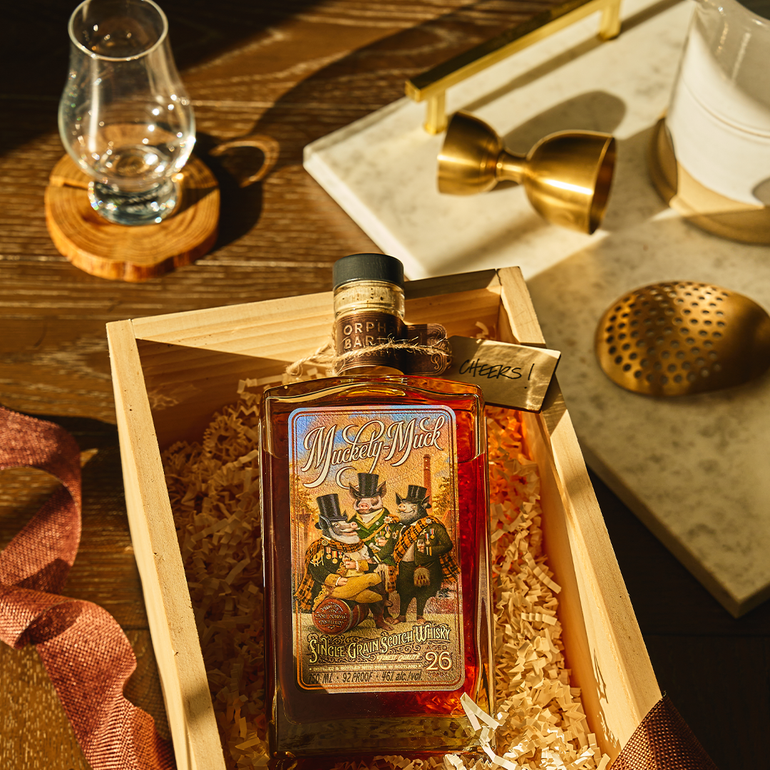 A bottle of Michter’s whiskey rests in a wooden box filled with packing straw, surrounded by a whiskey glass, jigger, strainer, and ribbon on a wooden surface.