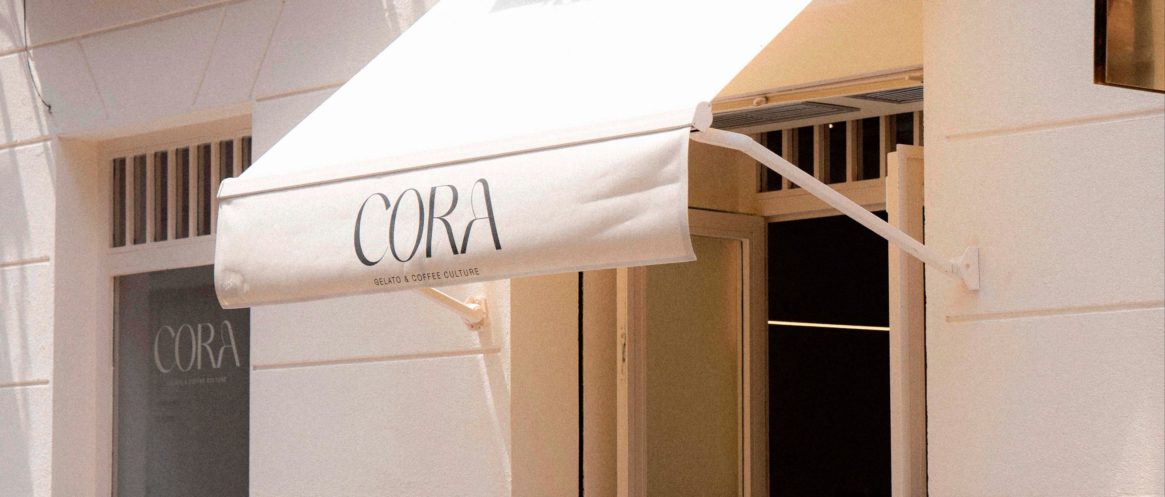 Toldo blanco con el nombre Cora y la frase “Gelato & Coffee Culture” en letras negras, sobre fachada clara de estética minimalista.