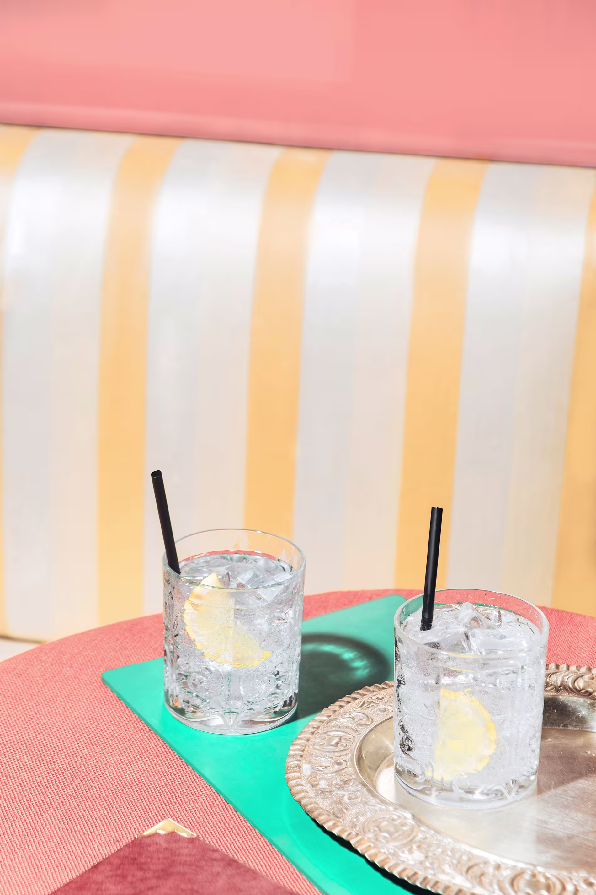Two glasses of clear beverage with lemon slices and black straws sit on a table, one on a silver tray, against a background of striped upholstery and pink wall.