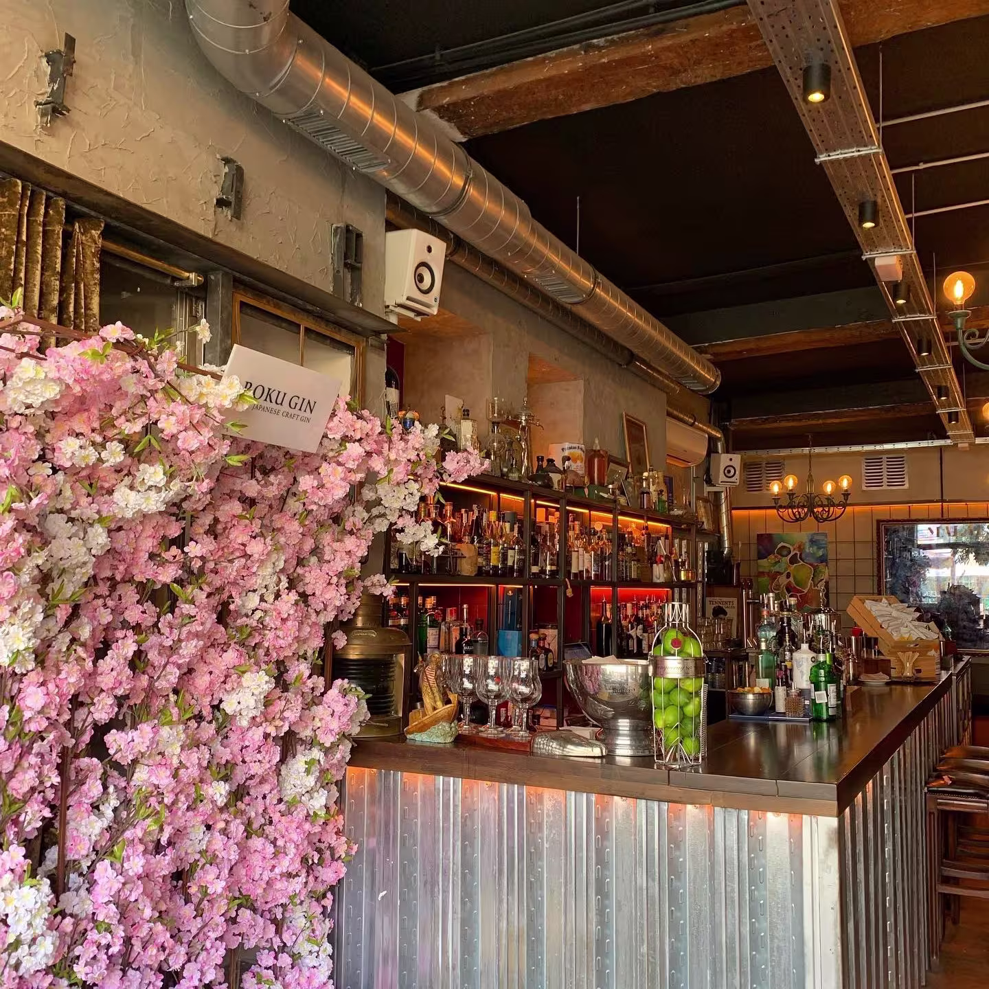 Barra metálica con estructura industrial, adornada con flores de cerezo artificiales, copas, botellas y limas, en un ambiente acogedor con luces cálidas y decoración colorida al fondo.
