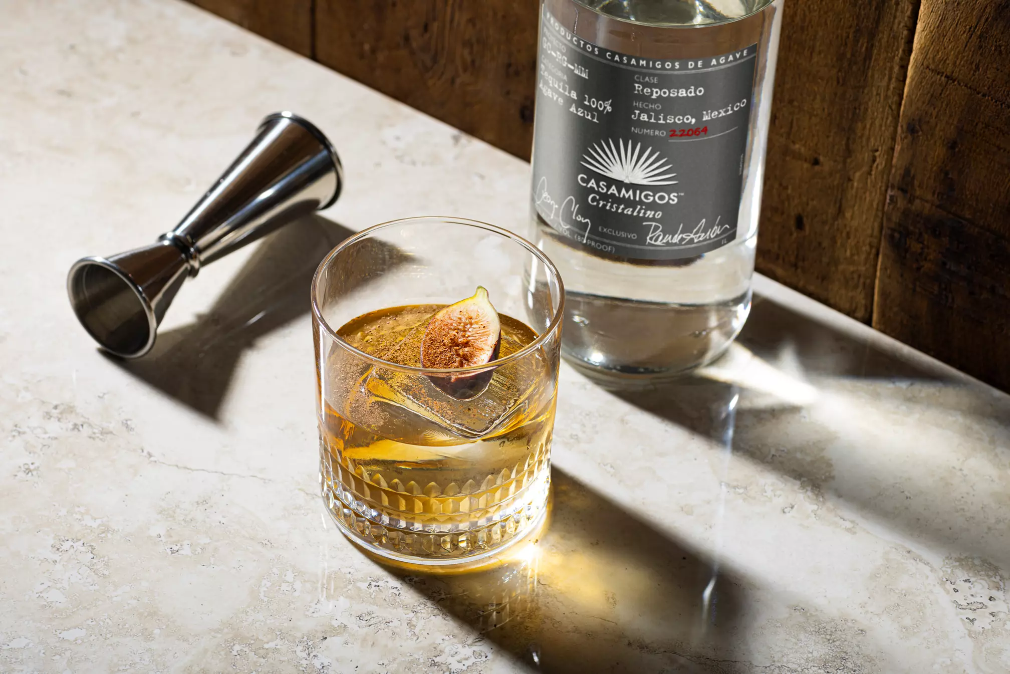 A glass of liquor with a fig garnish sits on a marble surface next to a bottle of Casamigos Cristalino tequila and a metal jigger.